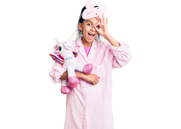 Menina afro-americana bonito vestindo máscara de sono e pijama segurando unicórnio de pelúcia rosa sorrindo feliz fazendo sinal ok com a mão no olho olhando através dos dedos  - Foto, Imagem