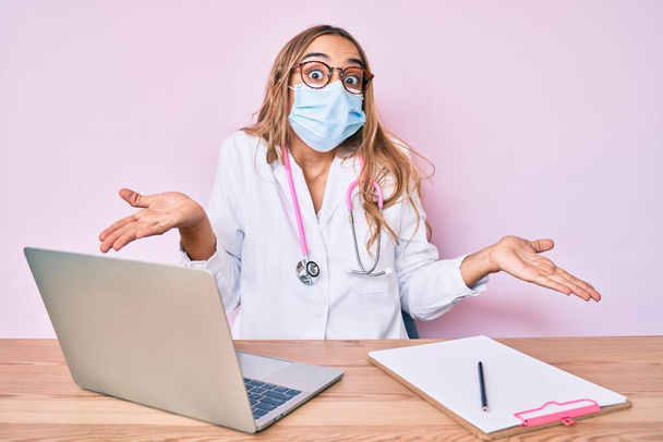Young beautiful blonde woman wearing doctor uniform and medical mask clueless and confused expression with arms and hands raised. doubt concept.  - Photo, Image
