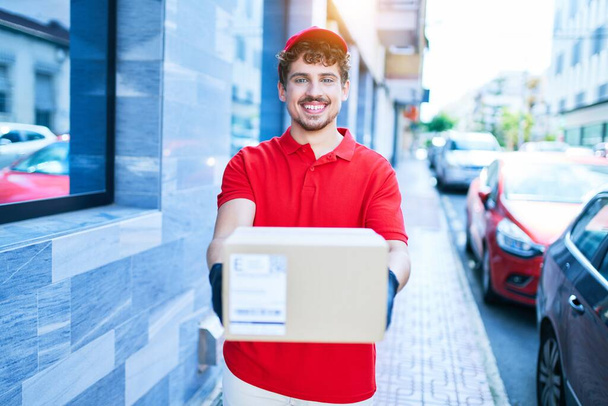 młody kaukaski dostawca człowiek uśmiechnięty szczęśliwy gospodarstwa karton pakiet spacery w mieście. - Zdjęcie, obraz