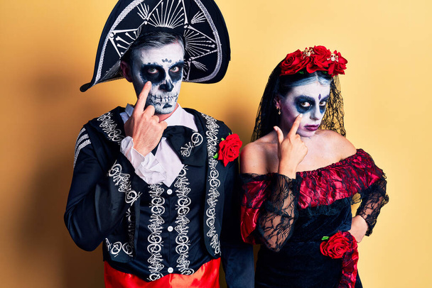 Young couple wearing mexican day of the dead costume over yellow pointing to the eye watching you gesture, suspicious expression  - Foto, Bild