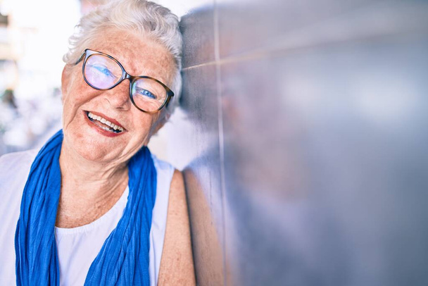 Anciana mujer mayor con el pelo gris sonriendo feliz apoyado en la pared al aire libre - Foto, imagen