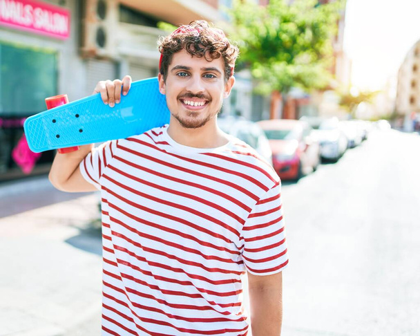Jovem homem patinador caucasiano sorrindo feliz segurando skate andando na rua da cidade. - Foto, Imagem