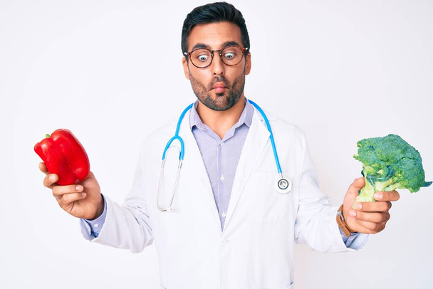 Young hispanic man wearing doctor stethoscope holding red pepper and broccoli making fish face with mouth and squinting eyes, crazy and comical.  - Photo, Image