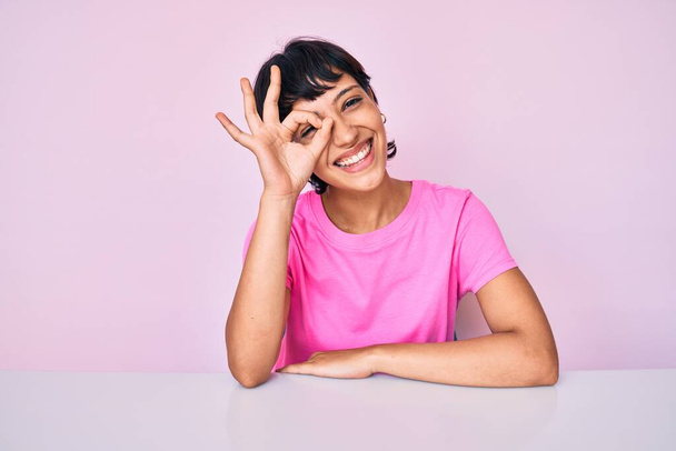 Belle femme brunettte portant des vêtements décontractés sur fond rose souriant heureux faisant signe ok avec la main sur les yeux regardant à travers les doigts  - Photo, image