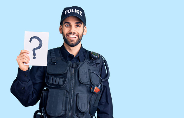 Junger gutaussehender Mann mit Bart, der Polizeiuniform trägt und ein Fragezeichen hinter sich hat, sieht positiv und glücklich stehend aus und lächelt mit einem selbstbewussten Lächeln, das Zähne zeigt  - Foto, Bild