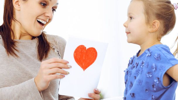 Mothers day concept. Child daughter congratulates mom and gives her postcard with red heart shape. Mum and girl happy smiling and hugging. Family fun and holiday - Valokuva, kuva