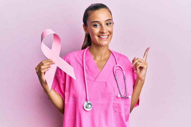 Mujer hispana joven sosteniendo cinta rosa de cáncer sonriendo feliz señalando con la mano y el dedo hacia un lado  - Foto, Imagen
