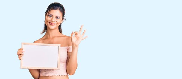 Jonge mooie vrouw met lege witte schoolbord doet ok teken met de vingers, glimlachend vriendelijk gebaar uitstekend symbool  - Foto, afbeelding