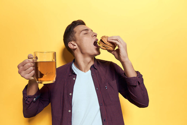 Χαρούμενος άνθρωπος με μια κούπα μπύρα και ένα χάμπουργκερ στα χέρια του δίαιτα τροφίμων τρόπο ζωής κίτρινο φόντο - Φωτογραφία, εικόνα