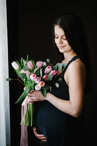 Pregnant woman hold bouquet of flowers in hands, girl embraces a round stomach belly near the window at home. Standing sideways. Maternity concept. Close up. nine months. Baby Shower. - Foto, afbeelding