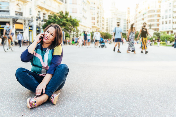 Esmer kadın, gündelik giysiler içinde Avrupa 'da bir meydanda insanlarla oturuyor ve cep telefonuyla konuşuyor. Boşluğu kopyala - Fotoğraf, Görsel