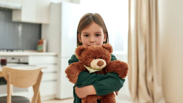 Ένα κοριτσάκι αγκαλιάζει ένα χνουδωτό καφέ αρκουδάκι κοιτάζοντας μια κάμερα. - Φωτογραφία, εικόνα