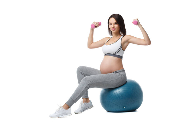 Slender athletic pregnant girl is engaged in fitness with dumbbells sitting on a ball isolated on a white background. - Фото, зображення