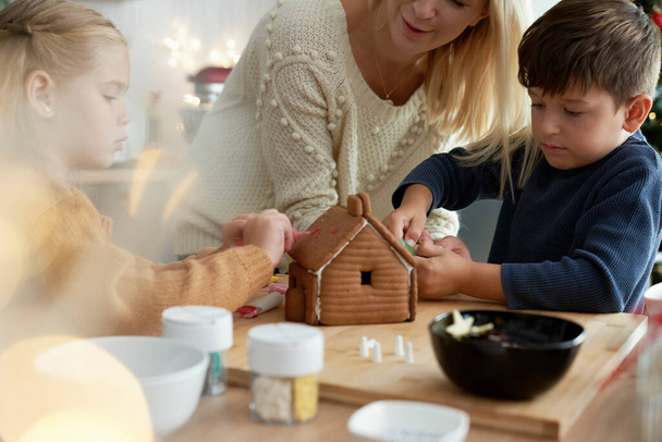 Children and mother spending time on decorating gingerbread house  - Photo, Image