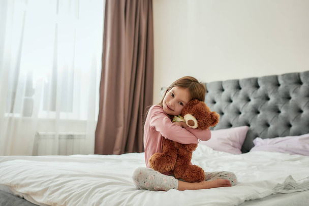 A cute small girl smiling and embracing a teddybear while sitting barefoot cross-legged on a bed at home - Foto, imagen