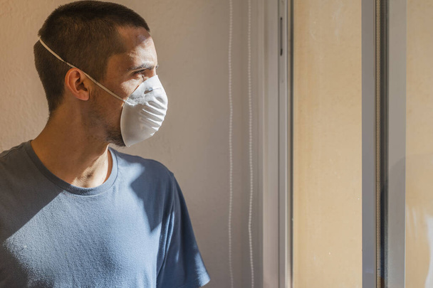 Koronavirus. Nemocný muž koronového viru, který se dívá oknem a nosí masku a zotavuje se z nemoci doma. Karanténa. Pacient izolován, aby se zabránilo infekci. Housi. Svoboda - Fotografie, Obrázek