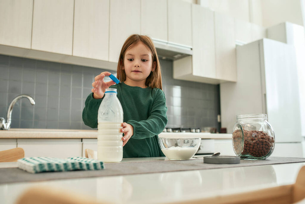 Holčička se chystá šukat uzávěr láhve mléka, když je v jasné kuchyni. - Fotografie, Obrázek