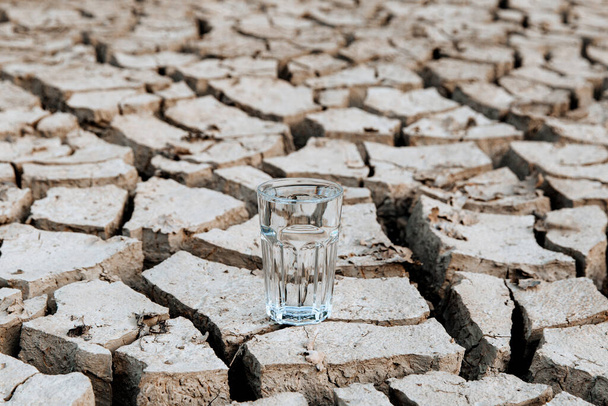 Egy átlátszó pohár tiszta ivóvíz áll a száraz, repedezett sivatagi föld közepén. Globális felmelegedés. Aszály és vízválság - Fotó, kép