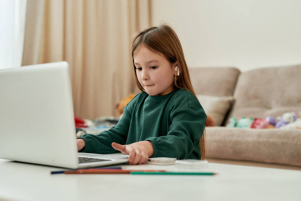 Una linda chica decidió hacer su hometask educación a distancia y mover un ordenador portátil en una mesa mientras estaba sentado solo en una habitación grande y luminosa - Foto, imagen