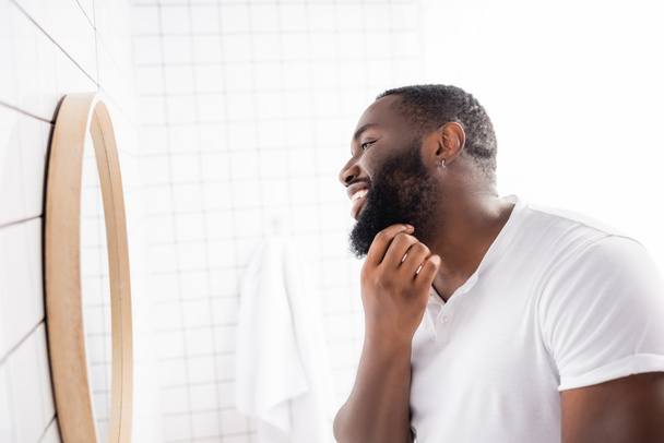 vista lateral del hombre afro-americano feliz mirando en el espejo y tocando la barba - Foto, imagen