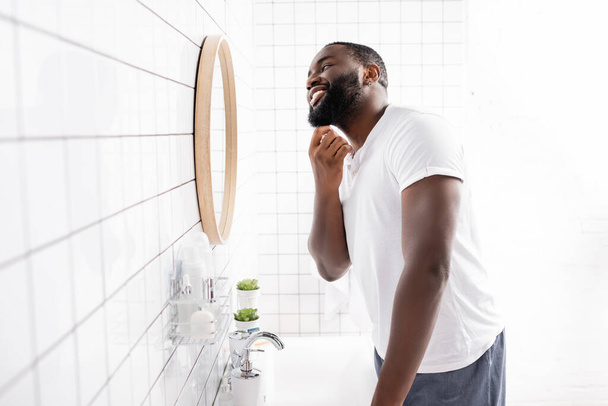 щасливий афроамериканський чоловік, дивлячись у дзеркало і торкаючись бороди у ванній
 - Фото, зображення