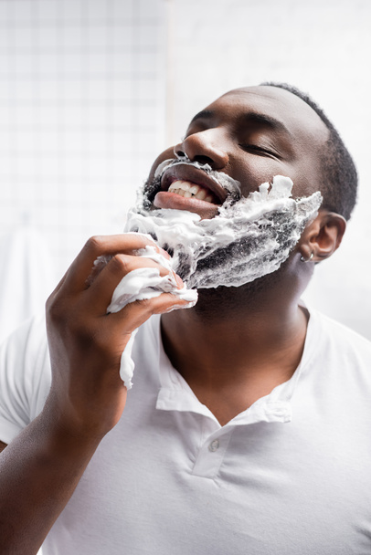 hombre afro-americano feliz con los ojos cerrados aplicando espuma de afeitar - Foto, imagen