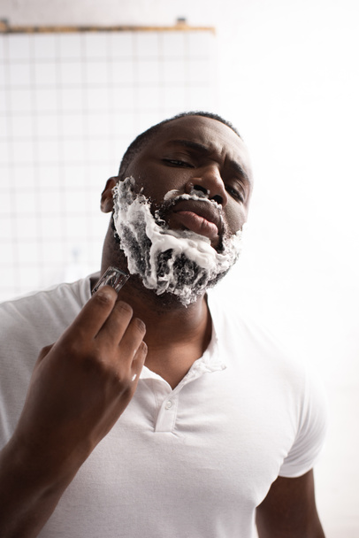 portrait of afro-american man with closed eyes shaving beard with razor  - Fotó, kép