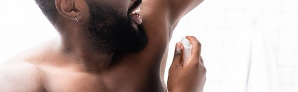plano panorámico del hombre afroamericano sonriente con la mano hacia arriba usando desodorante - Foto, Imagen