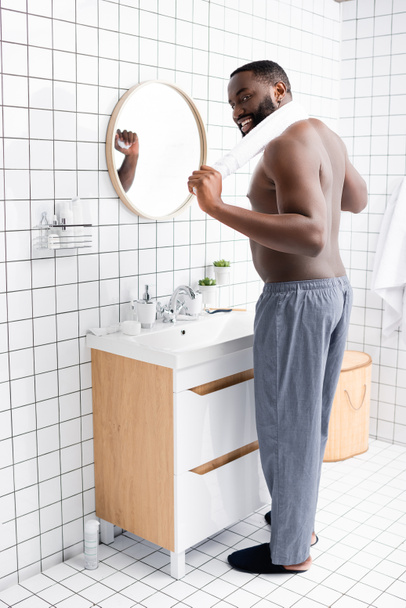 longitud completa del hombre afroamericano sonriente de pie en el baño con toalla detrás del cuello  - Foto, Imagen
