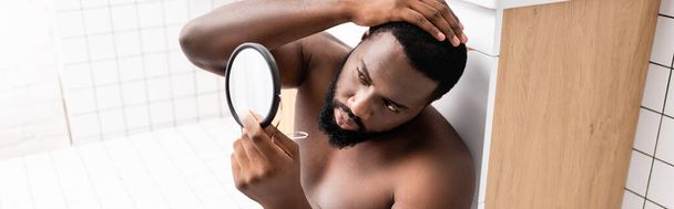 panoramic shot of afro-american man sitting on bathroom floor and fixing hair in small mirror - Photo, Image