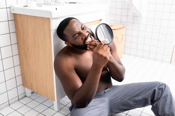 hombre afroamericano sentado en el piso del baño y mirando chicle en un pequeño espejo - Foto, Imagen