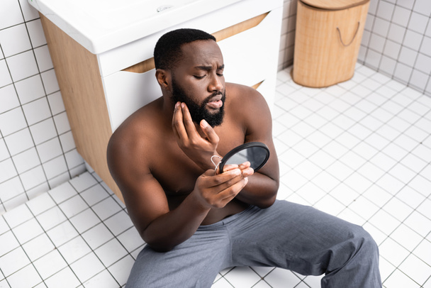 hombre afroamericano sentado en el suelo del baño y fijando el pelo en la barba - Foto, imagen