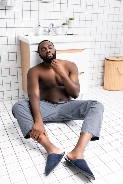 hombre afroamericano pensativo sentado en el suelo y tocando la barba  - Foto, Imagen
