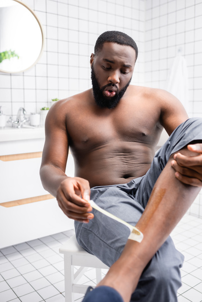 hombre afroamericano arrancando tiras de cera de la pierna - Foto, Imagen