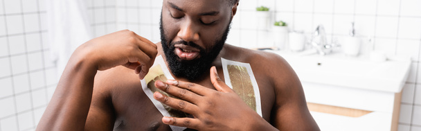 tiro panorâmico de homem afro-americano aplicando tiras de cera no peito  - Foto, Imagem
