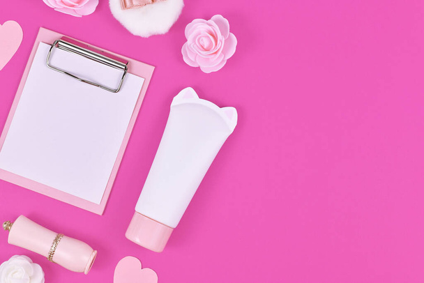 Flat lay with blank clipboard and various cute items like rose flowers, cream tube in shape of cat, lipstick and powder puff on side of pink background with empty copy space - Foto, Imagen