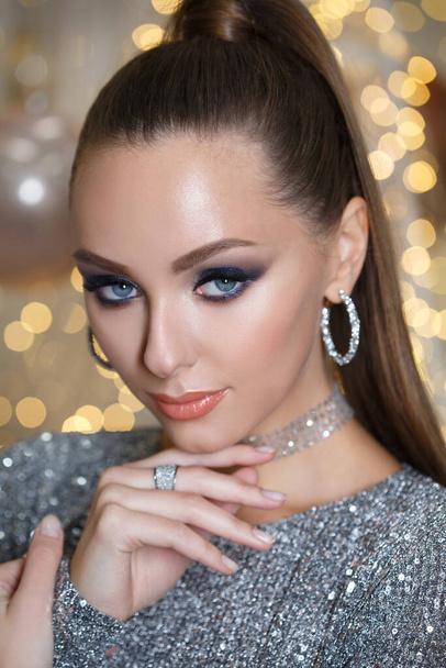 Beauty portrait of a beautiful girl with a silver shiny dress with evening make-up on a background of Christmas lights. - Фото, зображення