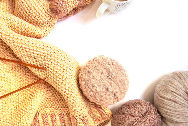 Balls of woolen yarn of nude colors and a knitted beige sweater, knitting needles, cup of coffee on a white background. Handmade woolen clothes. Traditional hobby. Copy space - Photo, Image