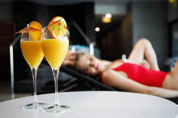 Zwei Weingläser mit Orangensaft, Scheiben und Sonnenschirmen auf dem Tisch. Vor dem Hintergrund liegt eine Frau im roten Badeanzug. Selektiver Fokus. Hintergrund verschwimmen. - Foto, Bild