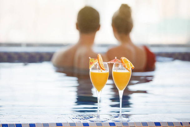 Due bicchieri di succo d'arancia con fette sullo sfondo di una coppia in piscina. Una donna e un uomo stanno nuotando nell'acqua blu. Concentrazione selettiva. Sfocatura sfondo. - Foto, immagini