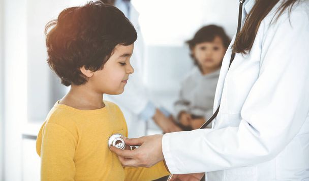 Woman-doctor examining a child patient by stethoscope in sunny clinik. Cute arab boy at physician appointment - Foto, Imagem