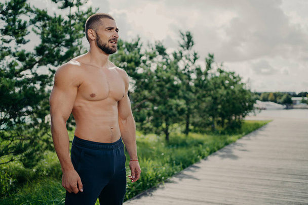 Confident sporty man bodybuilder has workout outdoor, looks thoughtfully into distance, wears shorts, has muscular body, strong biceps, stands over trees and sky background, shows strong torso - Photo, Image