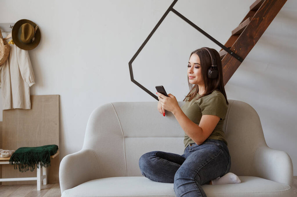 Cheerful young woman listening music in wireless headphones connected to smartphone in loft apartment - Photo, Image