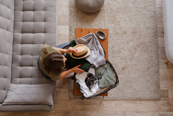 Προετοιμασία βαλίτσα για ένα ταξίδι διακοπών μετά το κλείδωμα. Νεαρή γυναίκα που ελέγχει ρούχα και πράγματα στις αποσκευές στον καναπέ στο σπίτι πριν από το ταξίδι - Φωτογραφία, εικόνα