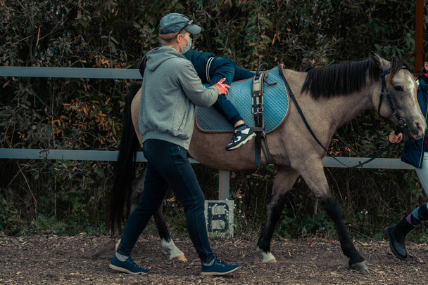 A little boy in turquoise overalls stroking an Icelandic pony horse with a funny forelock. The kid thanks the horse after hippotherapy. The instructor uses a mask during quarantine - Foto, afbeelding