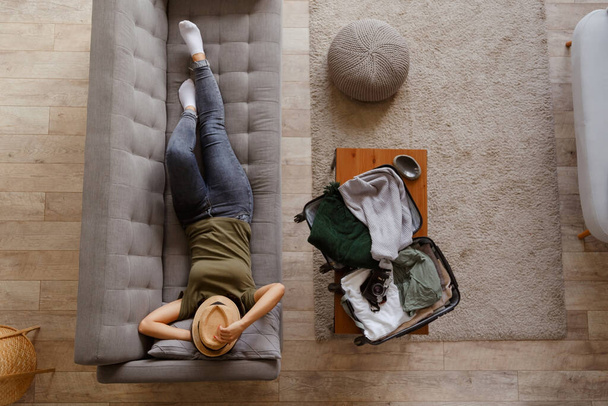 Young enthusiastic woman is relaxing on the couch at home before the upcoming trip after a long quarantine. View from above. A suitcase with things on the coffee table - Фото, изображение