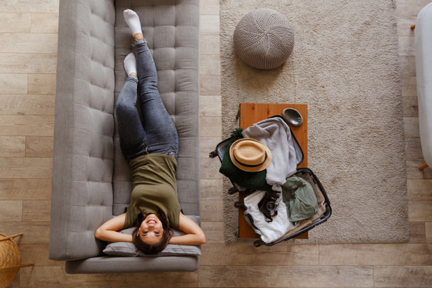 Young enthusiastic woman is relaxing on the couch at home before the upcoming trip after a long quarantine. View from above. A suitcase with things on the coffee table - Valokuva, kuva
