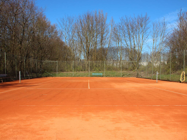 Un court de tennis vide en argile rouge en été. Il n'y a pas de filet, et la cour est entourée d'une clôture métallique avec des arbres en arrière-plan. - Photo, image