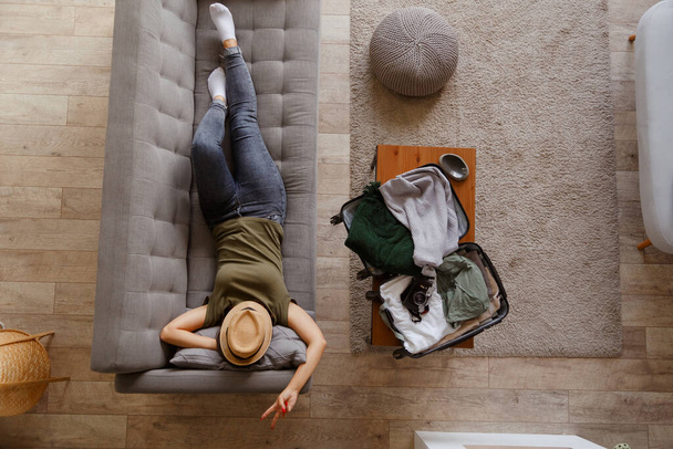 Молодая восторженная женщина отдыхает дома на диване перед предстоящей поездкой после длительного карантина. Вид сверху. Чемодан с вещами на журнальном столике - Фото, изображение