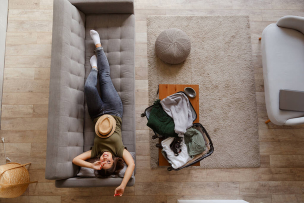 Nuori innostunut nainen rentoutuu sohvalla kotona ennen tulevaa matkaa pitkän karanteenin jälkeen. Näkymä ylhäältä. Matkalaukku, jossa on tavaraa sohvapöydällä. - Valokuva, kuva